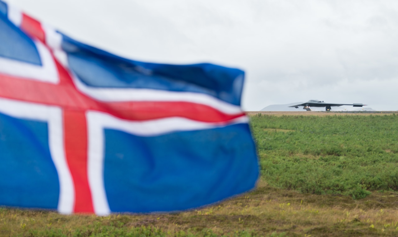 The flag of Iceland waves in the foreground. In the background, an airplane lands on a runway.
