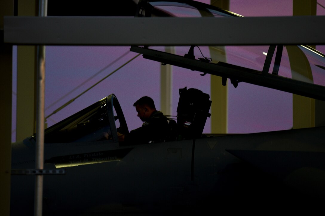 An Air Force pilot, shown in silhouette, sits in an open cockpit in a hangar-type structure, against a purple sky.