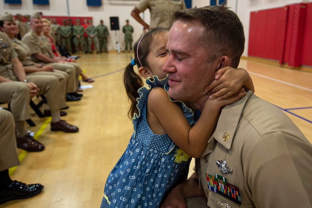 A girl kisses her father as he kneels on a gym floor, in front of an audience.