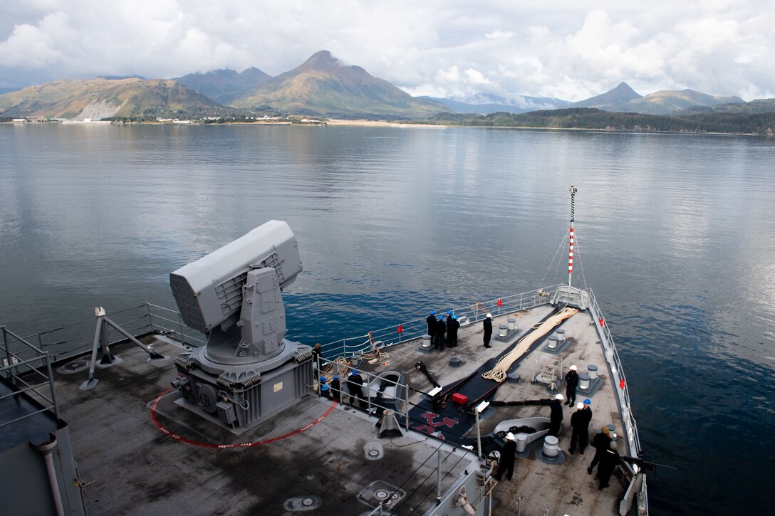 The front of a military ship heads across placid water to a mountainous coastline.