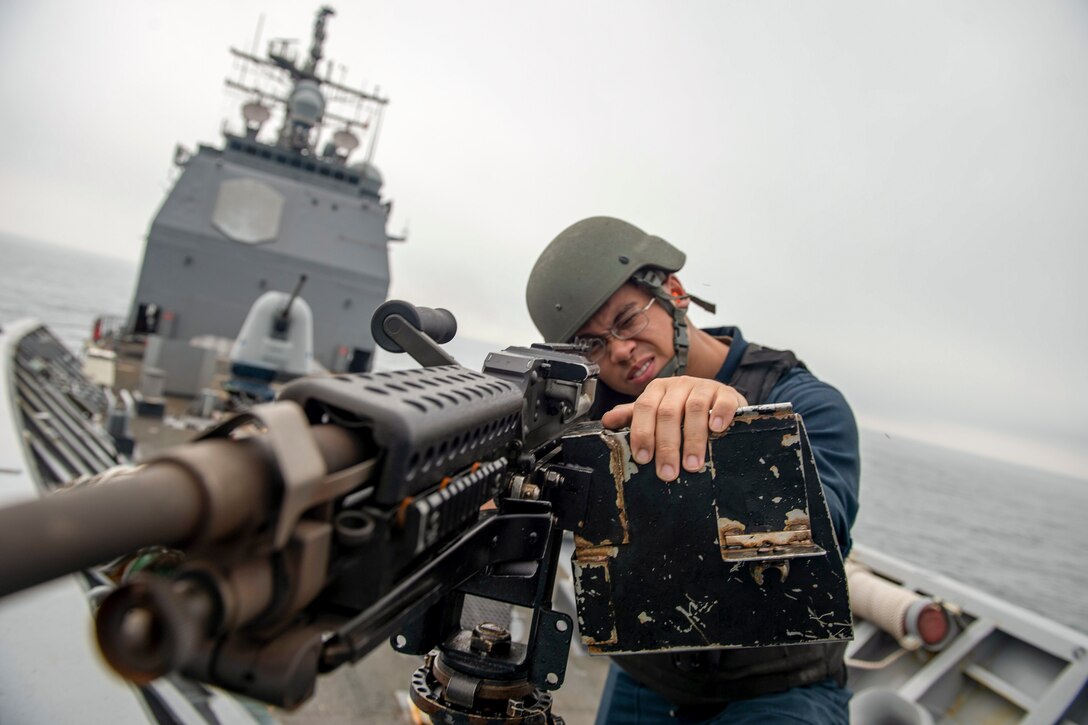 A sailor squints through the scope of a machine gun on a ship's forecastle.