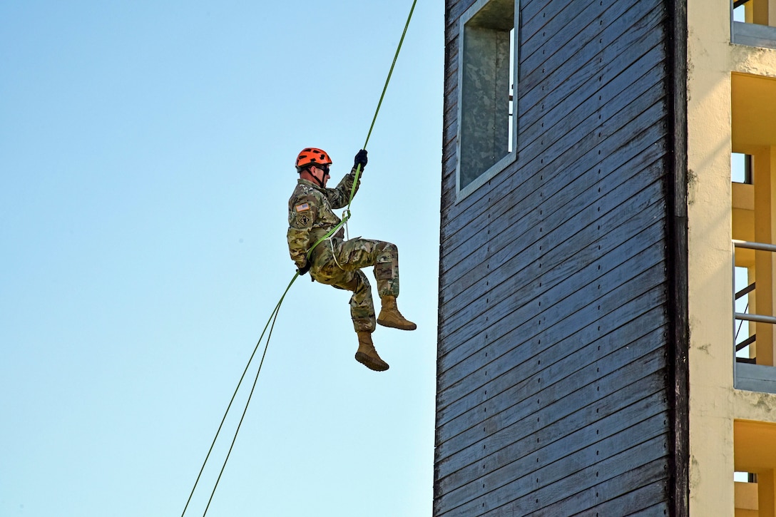 A soldier hangs in midair while rappelling from a tower.
