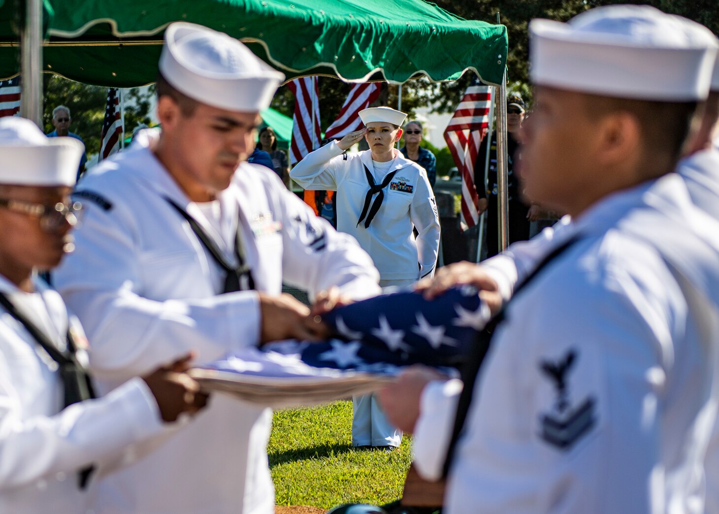 A sailor salutes while other sailors fold a flag.