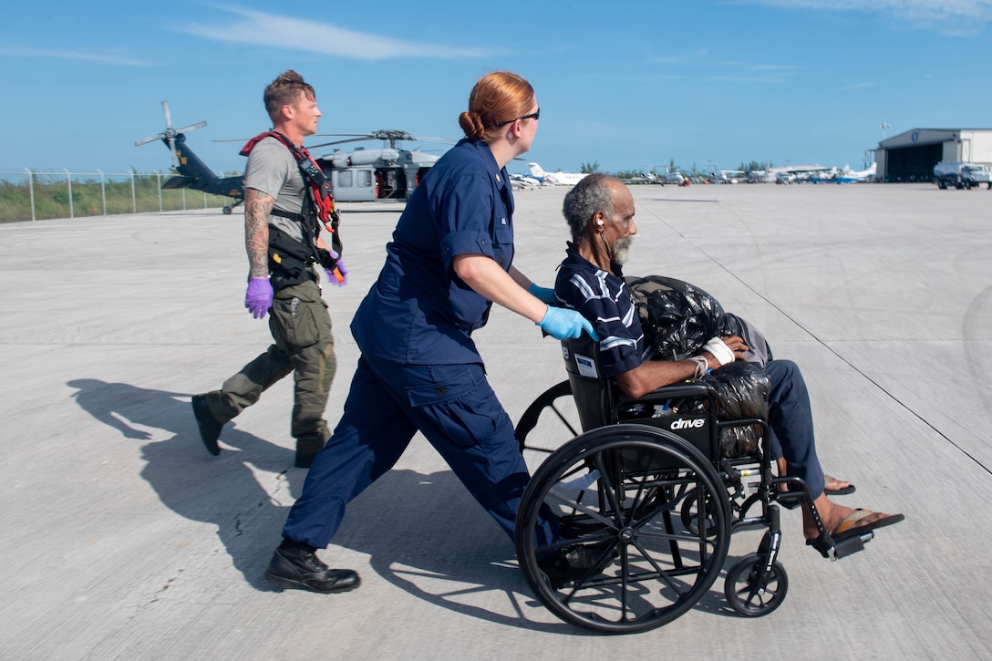 A service member pushes a man in a wheelchair on a flightline as another service member walks beside her.