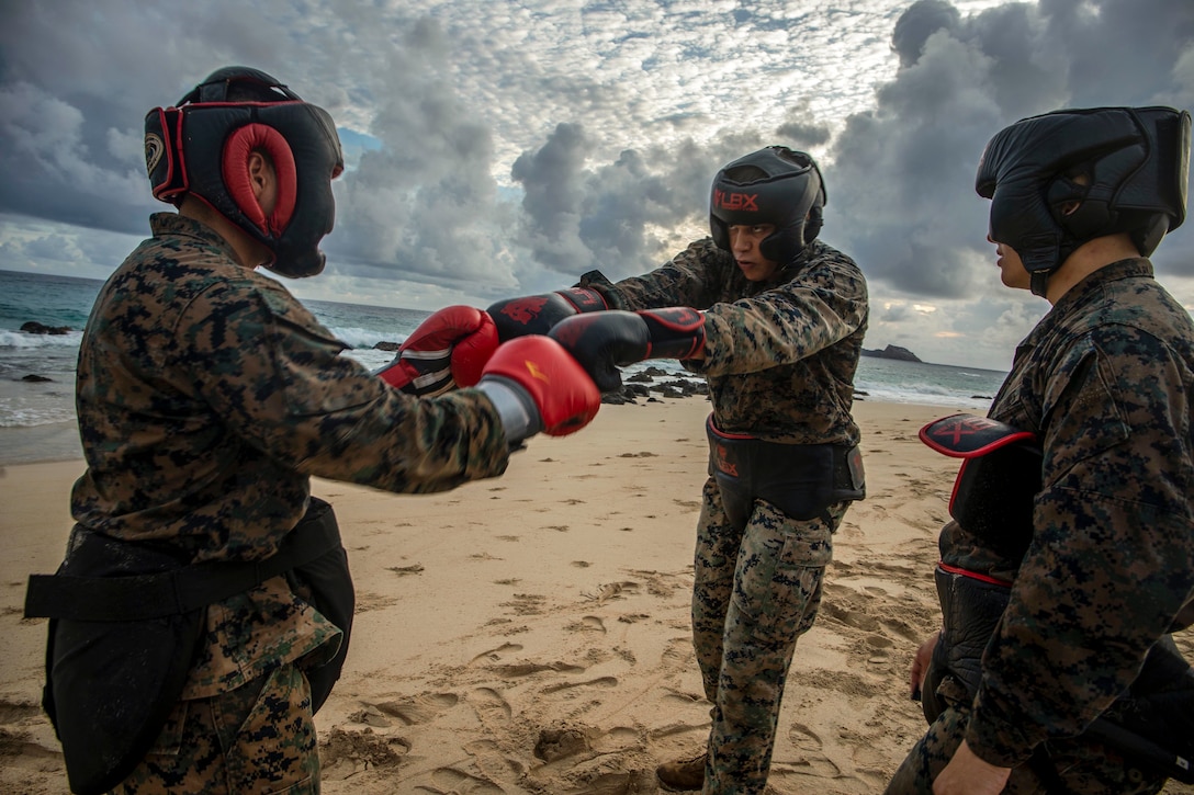 Two Marines tap boxing gloves while standing facing each other on a beach, as a third observes.