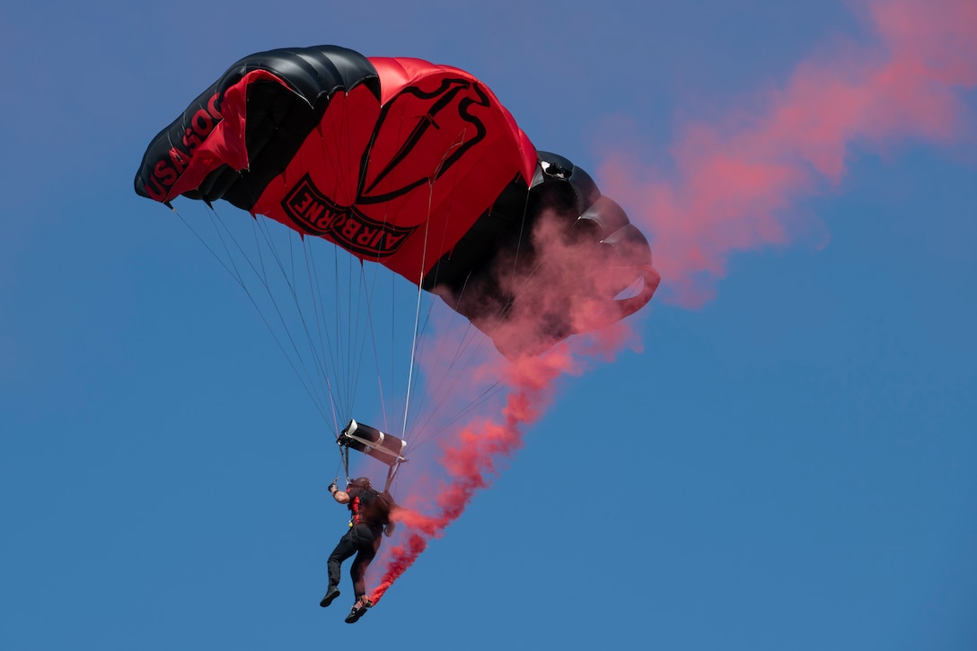 A parachutist prepares to land with a trail of red smoke behind him.