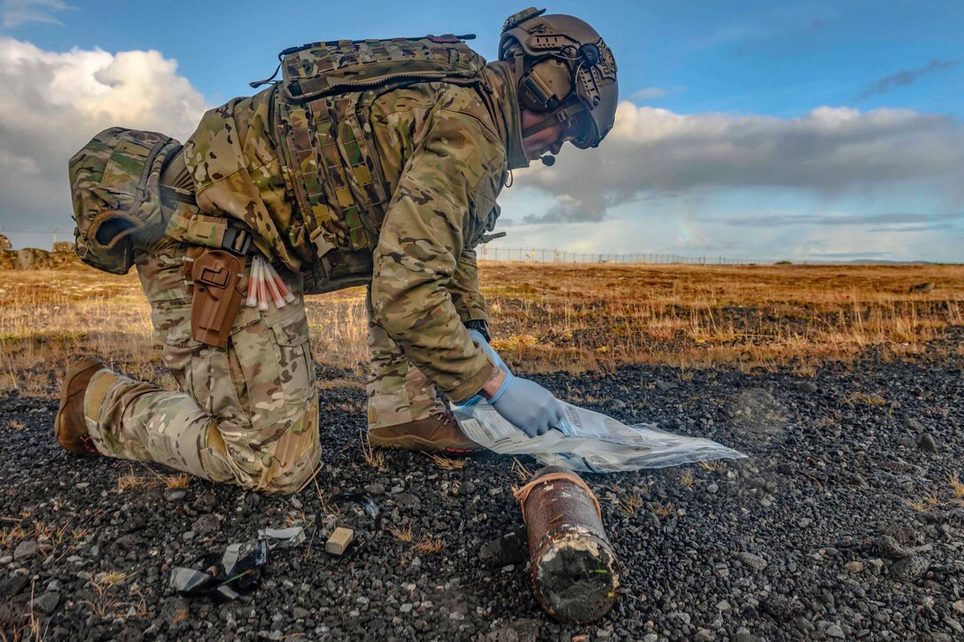 A sailor kneels on gravelly terrain near a mock bomb.