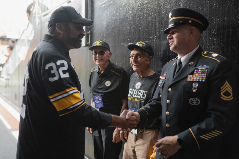 A former football player wearing a number 32 jersey shakes hands with a man in a military uniform as two elderly men look on.