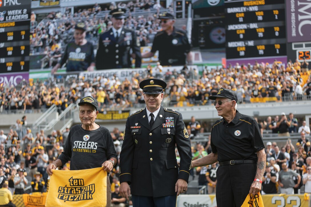 Two elderly men stand on a football field holding yellow banners; they are wearing caps with the words “World War II veteran.” In the background, the bleachers are filled with spectators; above the crowd, the men can be seen on a giant video screen. Between the elderly men stands in younger man wearing an Army uniform.