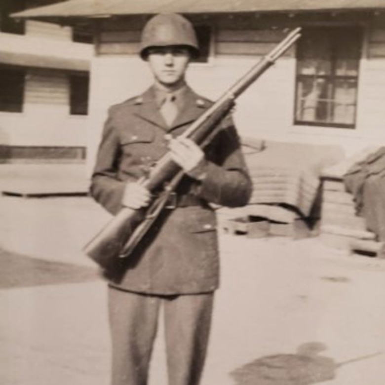 A young man dressed in an Army uniform poses hold up his rifle for a photo.