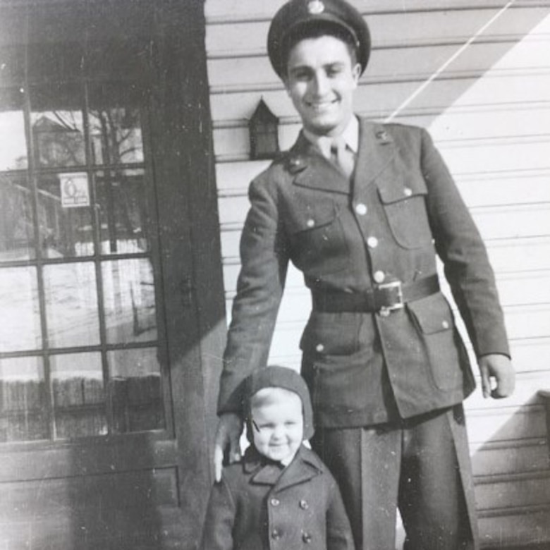 Man in an Army uniform smiles as he stands next to a small child, who is wearing a hat and coat