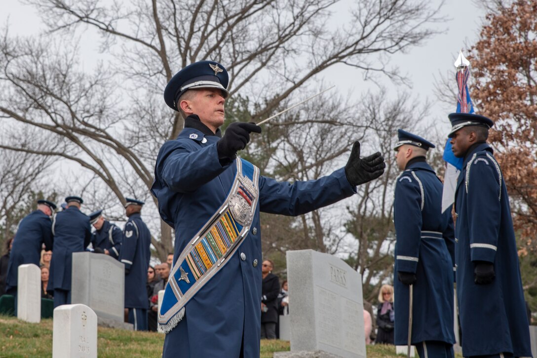 An airman in a dress uniform and winter coat holds his hands up to conduct a band. Two men stand in formation facing him. Several mourners are in the background near gravestones.