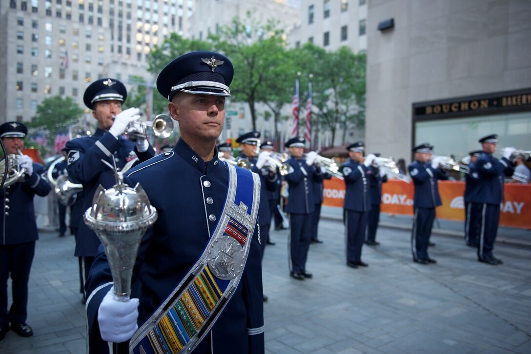 An airman wearing a formal hat, sash and mace stands in the foreground while several other airmen play brass instruments  in the background. Two U.S. flags are in the background.