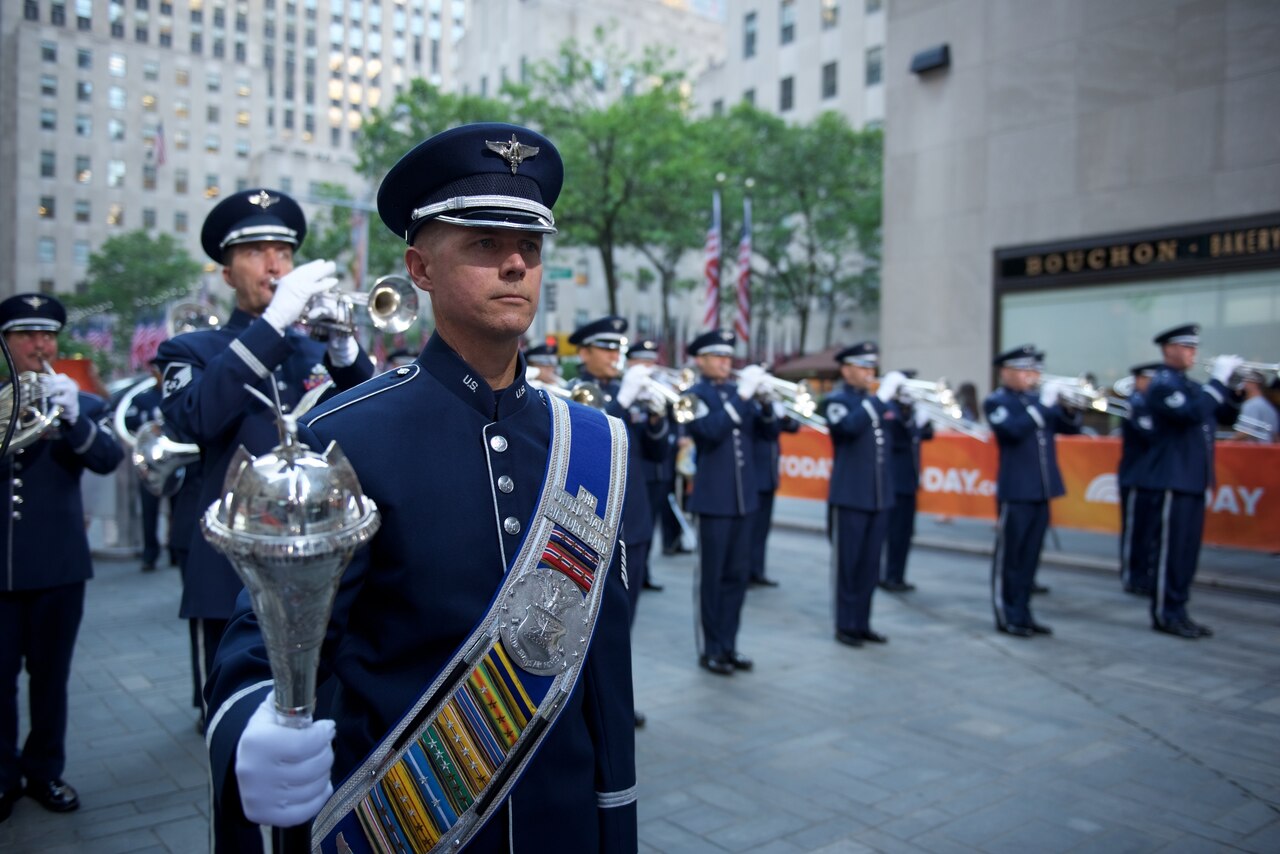 An airman wearing a formal hat, sash and mace stands in the foreground while several other airmen play brass instruments  in the background. Two U.S. flags are in the background.