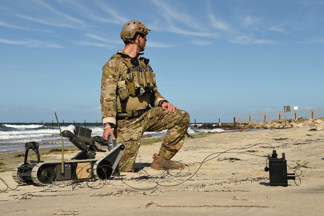 A man kneels on a beach with a small robotic device next to him.