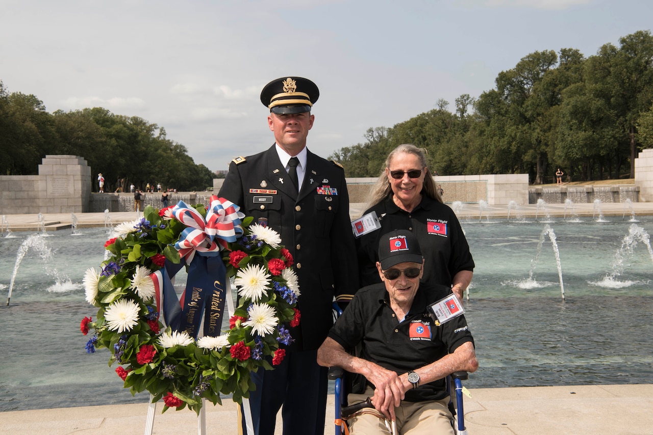A man in a wheelchair, a man in a military uniform and a woman stand next to a wreath at the World War II Memorial.