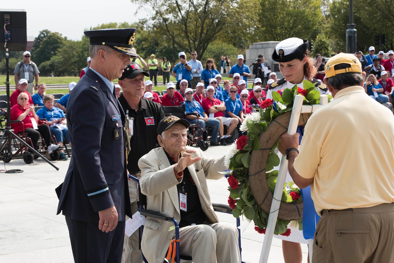 A man in a wheelchair helps place a wreath at an outdoor event.  He is accompanied by two military personnel and a man behind him pushing his chair.