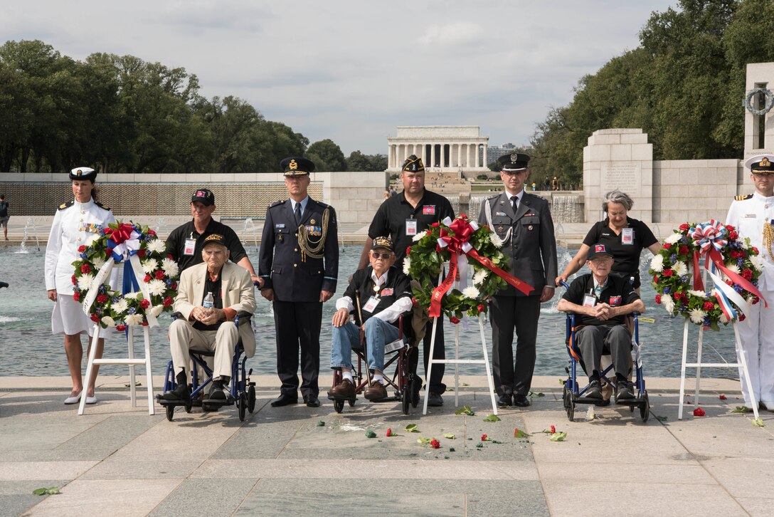 Three men in wheelchairs sit next to wreaths at an outdoor location. They are accompanied by both men and women in military uniforms. In the background are portions of the World War II Memorial and the Lincoln Memorial.