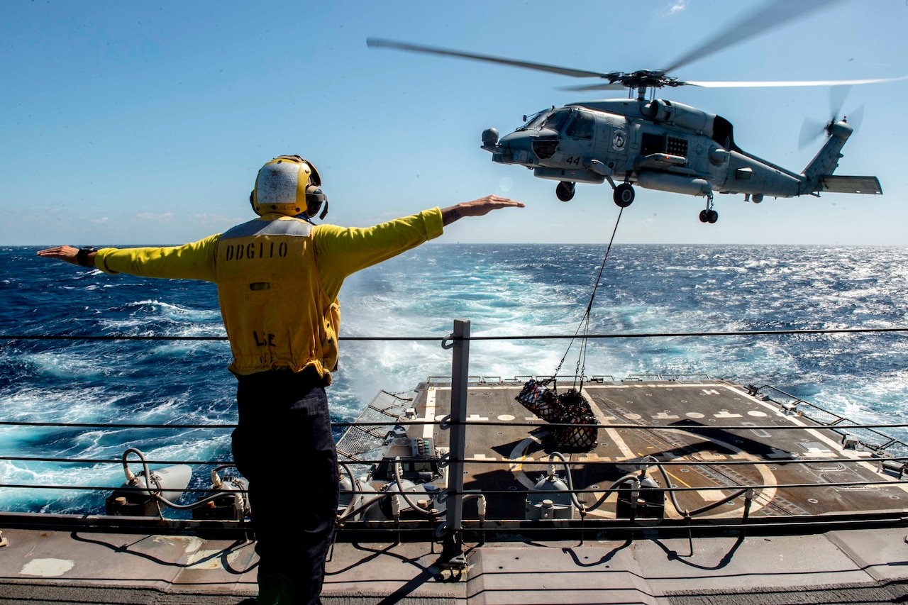 A sailor hold his arms out as a helicopter hovers above a military ship.