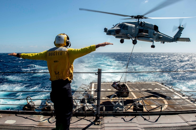 A sailor hold his arms out as a helicopter hovers above a military ship.