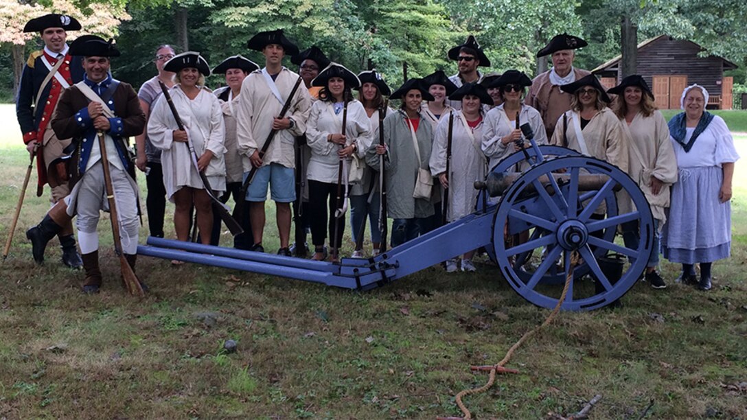 A line of students and teachers in Revolutionary War costumes pose behind a cannon