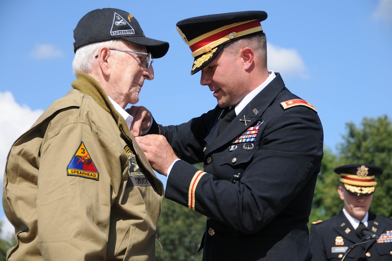 A man in a military uniform pins a medal to the lapel of another man in an outdoor location.