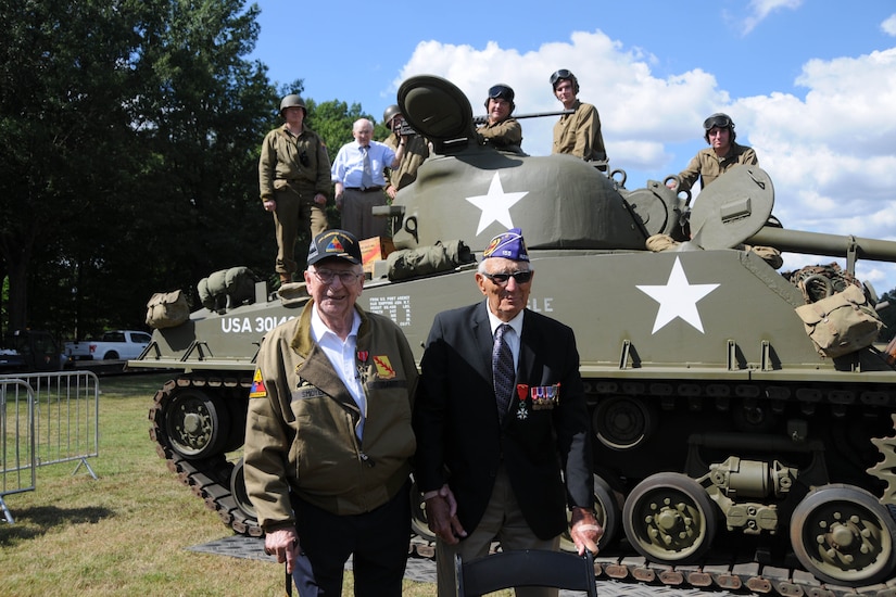 Two men stand in front of a large military tank.