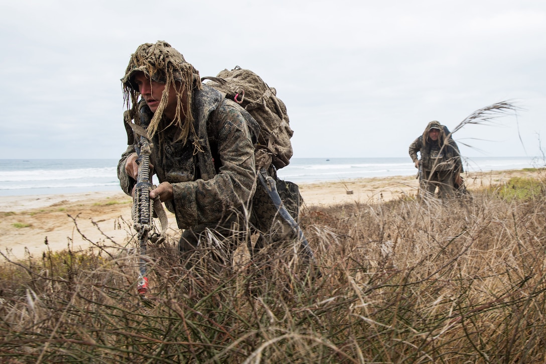 Two service members crouch and move forward on a beach.