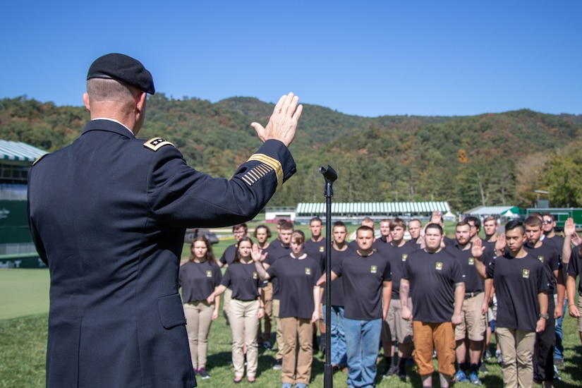 Older Army soldier raises his hand in front of large group of young teenagers in the middle of the valley.