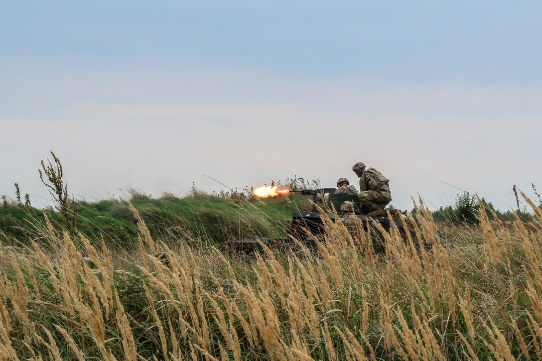 Soldiers fire a weapon from a vehicle in a field.