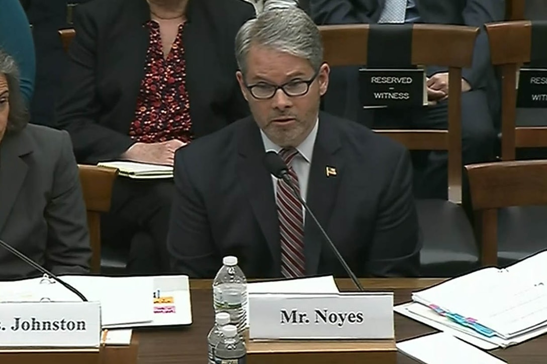 Man in suit sits in front of a microphone at a hearing.