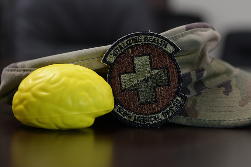 A brain-shaped stress ball and a 628th Medical Operations Squadron patch are displayed in front of an operational camouflage pattern cap at the mental health clinic Aug. 27, 2019, at Joint Base Charleston, S.C. Air Force medical technicians help ensure the medical readiness, both physically and mentally, of over 200,000 Airmen around the world.