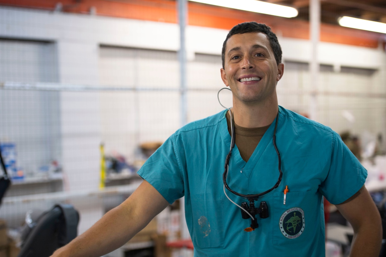 A Navy dentist wearing scrubs and stethoscope stands in a room and smiles.