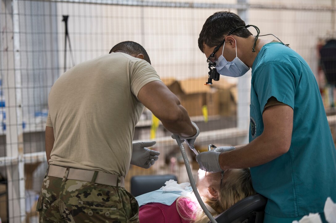 Two military dental professionals lean over a patient in a chair while performing work.