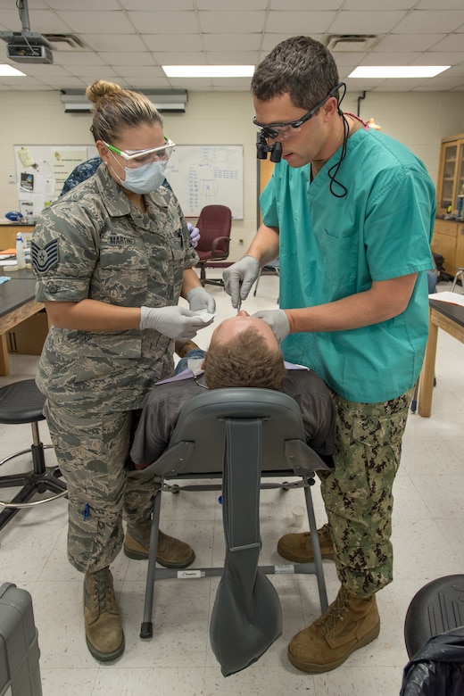 An Air Force dentist and Navy dentist stand on either side of a patient while performing a procedure.