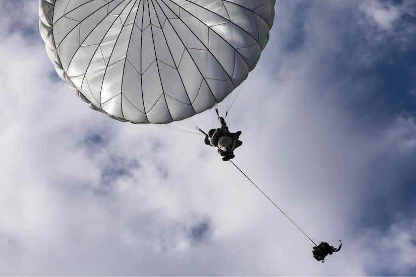 A paratrooper descends via parachute above his rucksack.