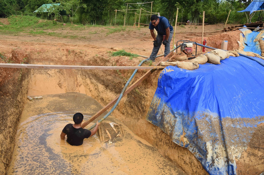 A service member stands in a muddy trench while another looks on.