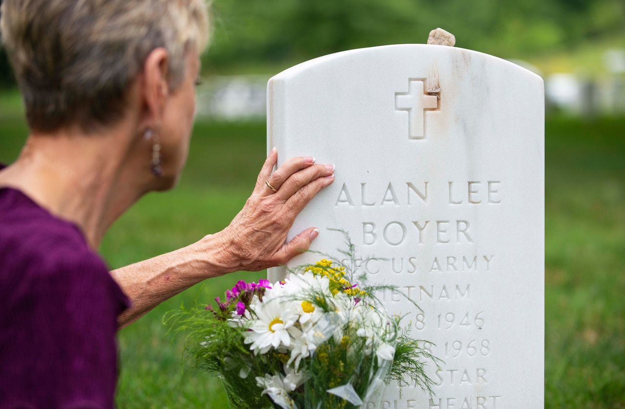 A woman touches a headstone with flowers at the base.
