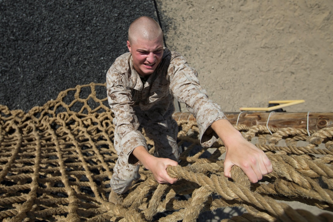 A Marine Corps recruit grimaces as he climbs up a cargo net.