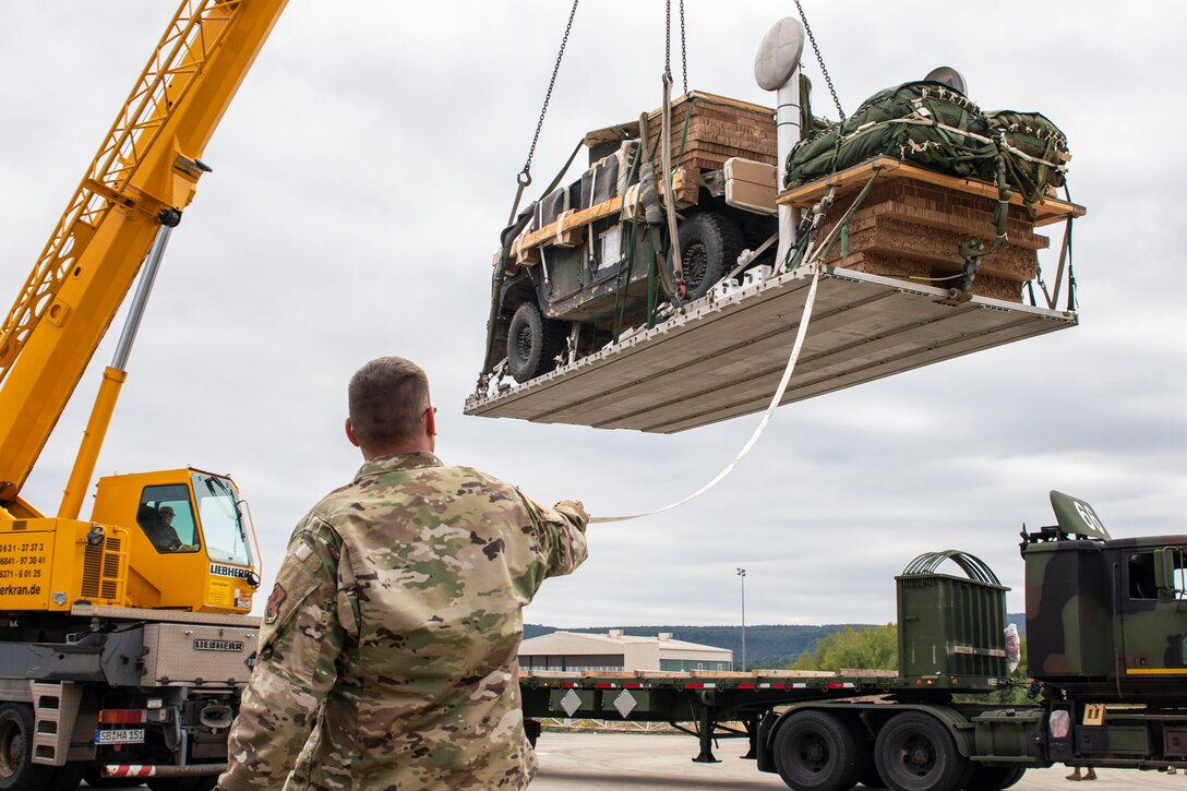 An airman holds onto a rope that is hanging from a platform that a large construction vehicle is carrying.
