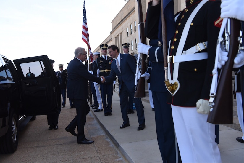 Men in suits shake hands while flanked by service members in dress uniforms.