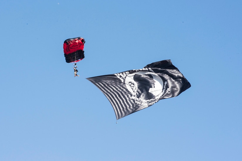 A service member descends through the air with a parachute and a flag,