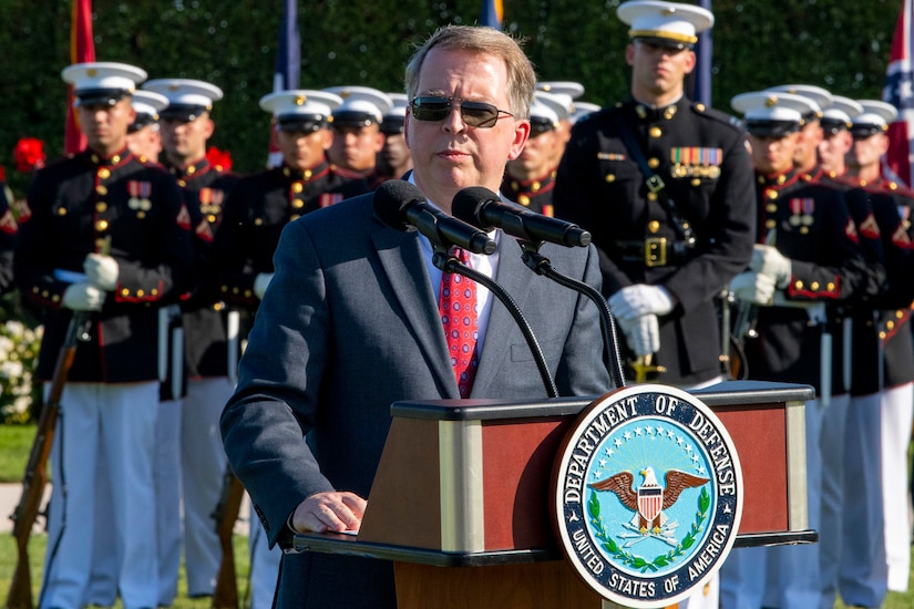 A man stands at a lectern.  Behind him are dozens of uniformed military personnel in formation.