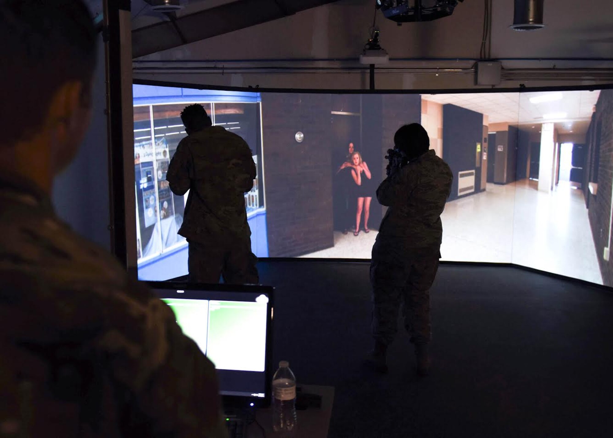 An Airman instructor watches two Airmen point inert firearms at a screen during an active school shooter simulation which shows a shooter holding a gun to a teenager's head.