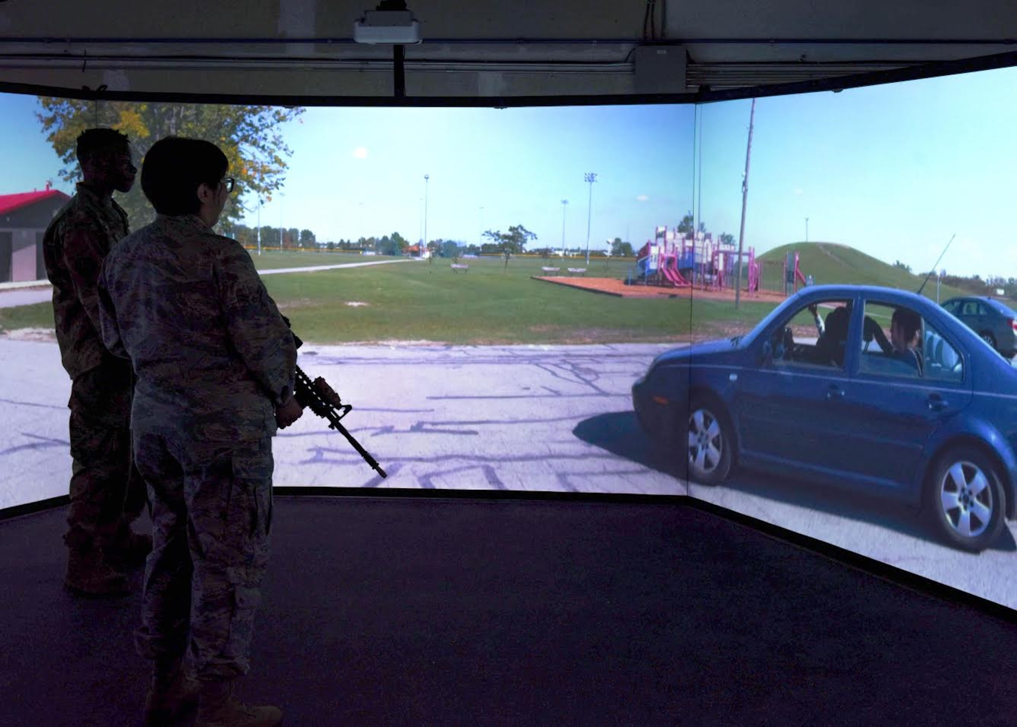 Two Airmen stand to the left of a screen, holding inert firearms, during a noise complaint simulation and speak to a car full of indivduals.