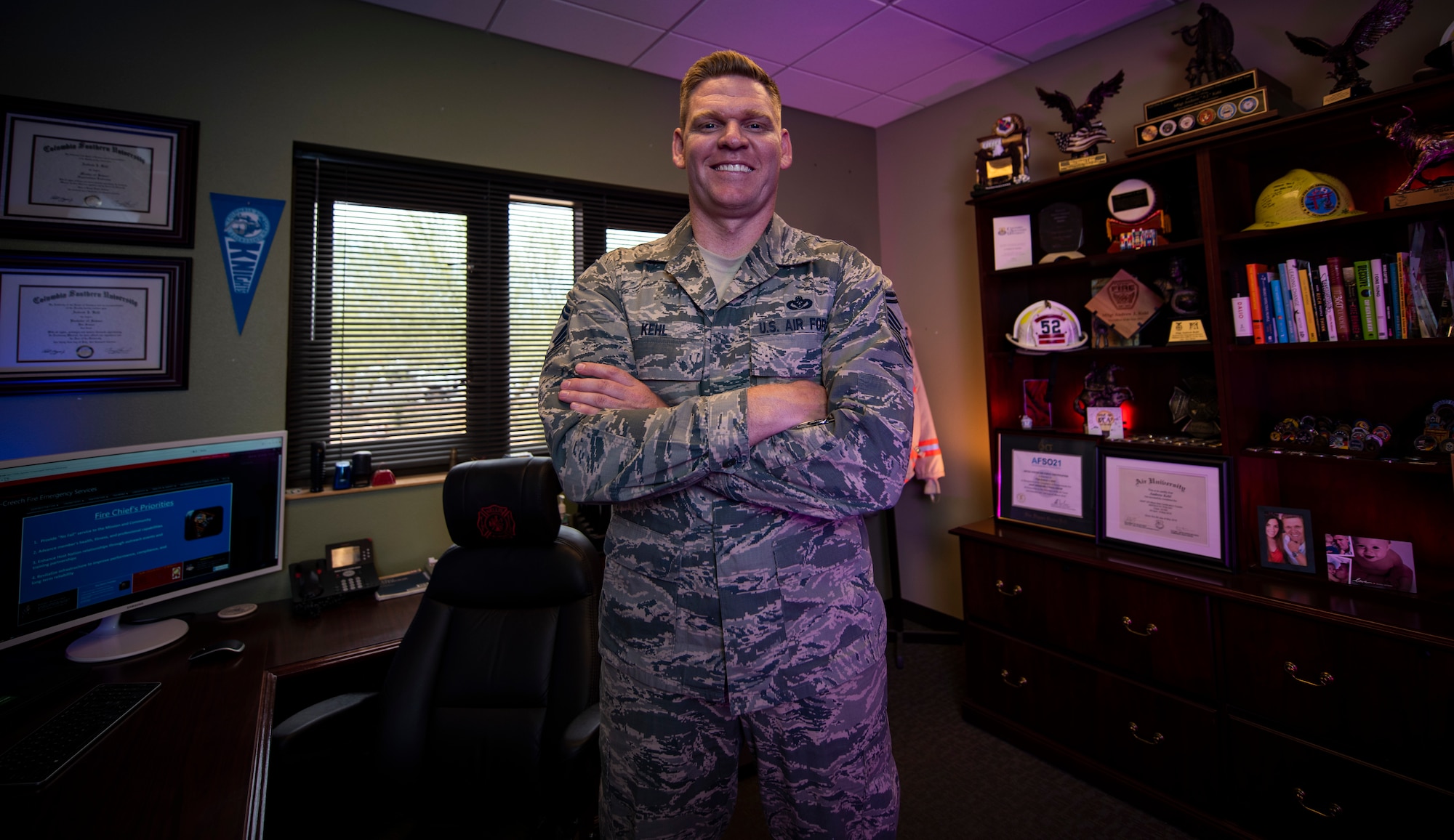 An Airman stands in the middle of his office and smiles for a photo.