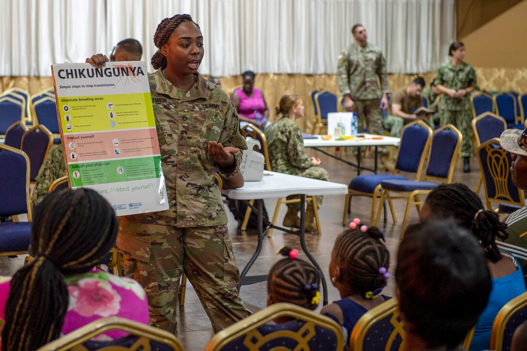 A soldier stands with poster and speaks to a group of kids and adults seated in chairs.