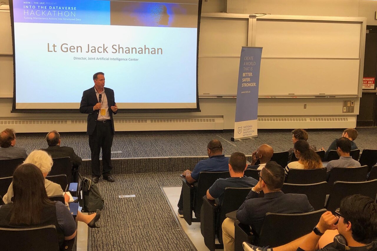 A man holding a microphone stands in front of a projection screen that reads “Lt. Gen. Jack Shanahan.”