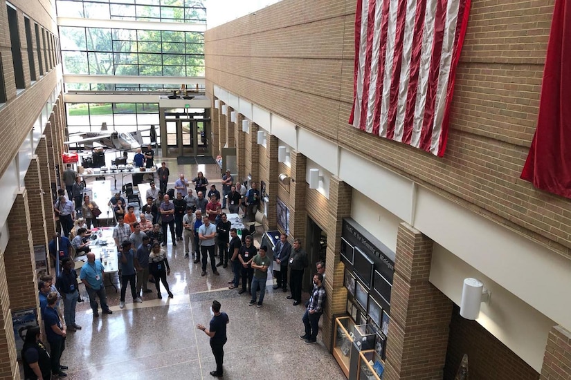 An overhead view of people gathered in a long, narrow sunlit foyer.