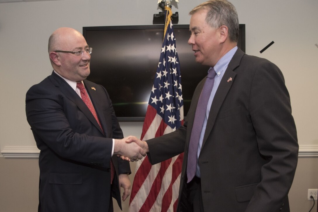 Two men shake hands in an office with a U.S. flag behind them.